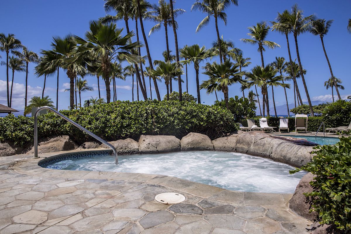 A serene outdoor hot tub surrounded by greenery and palm trees, with lounge chairs and the ocean in the background under a clear blue sky.