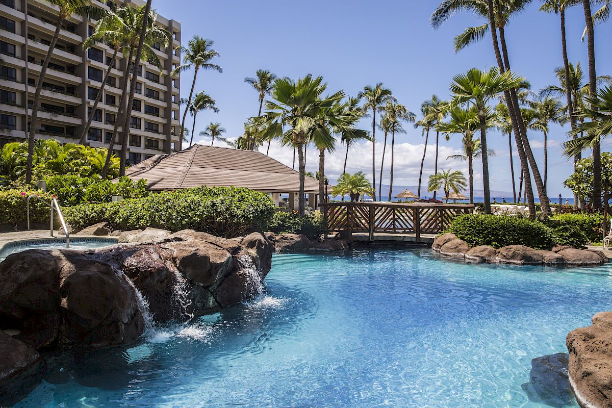 A tropical resort scene featuring a swimming pool, palm trees, rock waterfall, and a bridge, with a multistory building in the background.