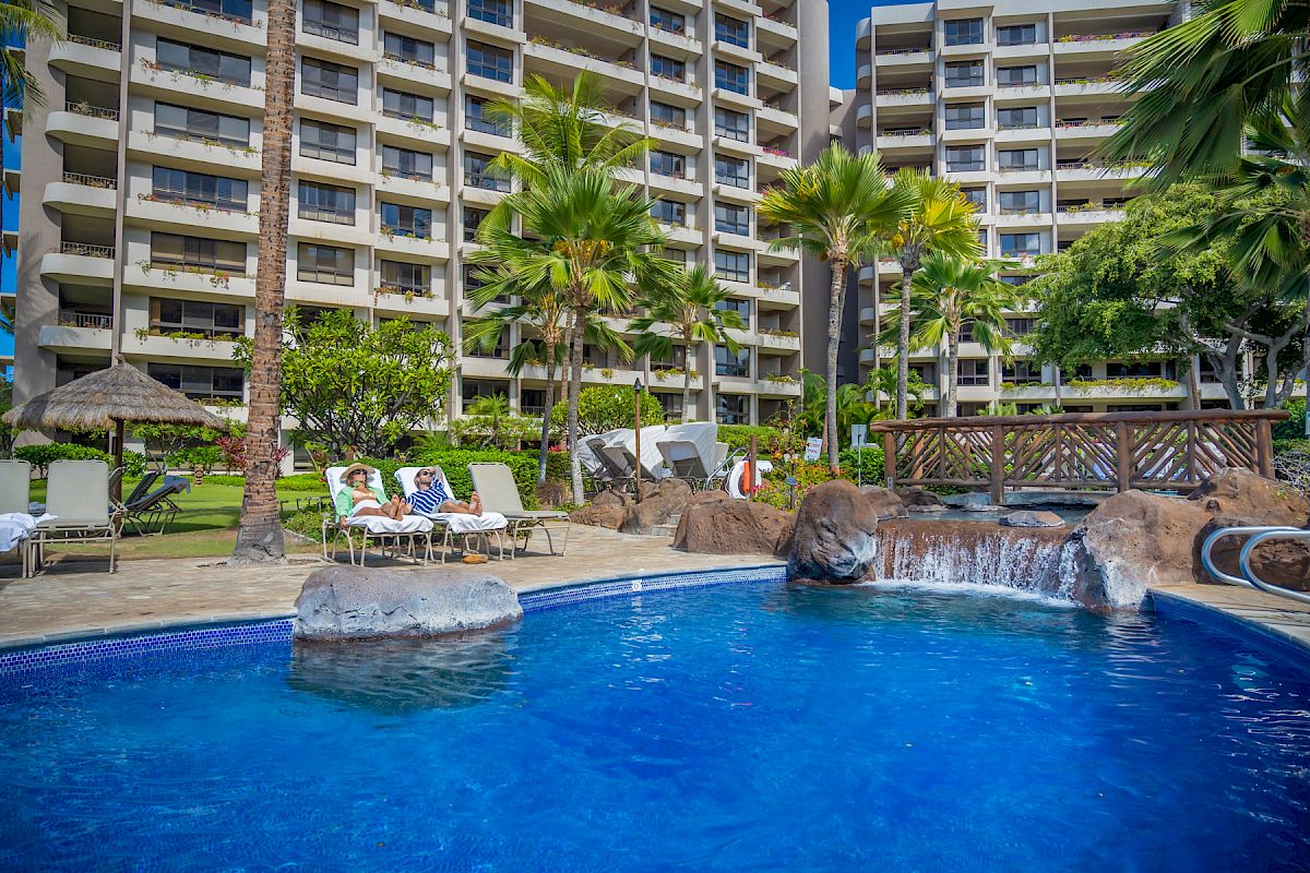 A swimming pool with a waterfall feature is in the foreground, surrounded by tall palm trees and lounge chairs in front of a multi-story hotel.