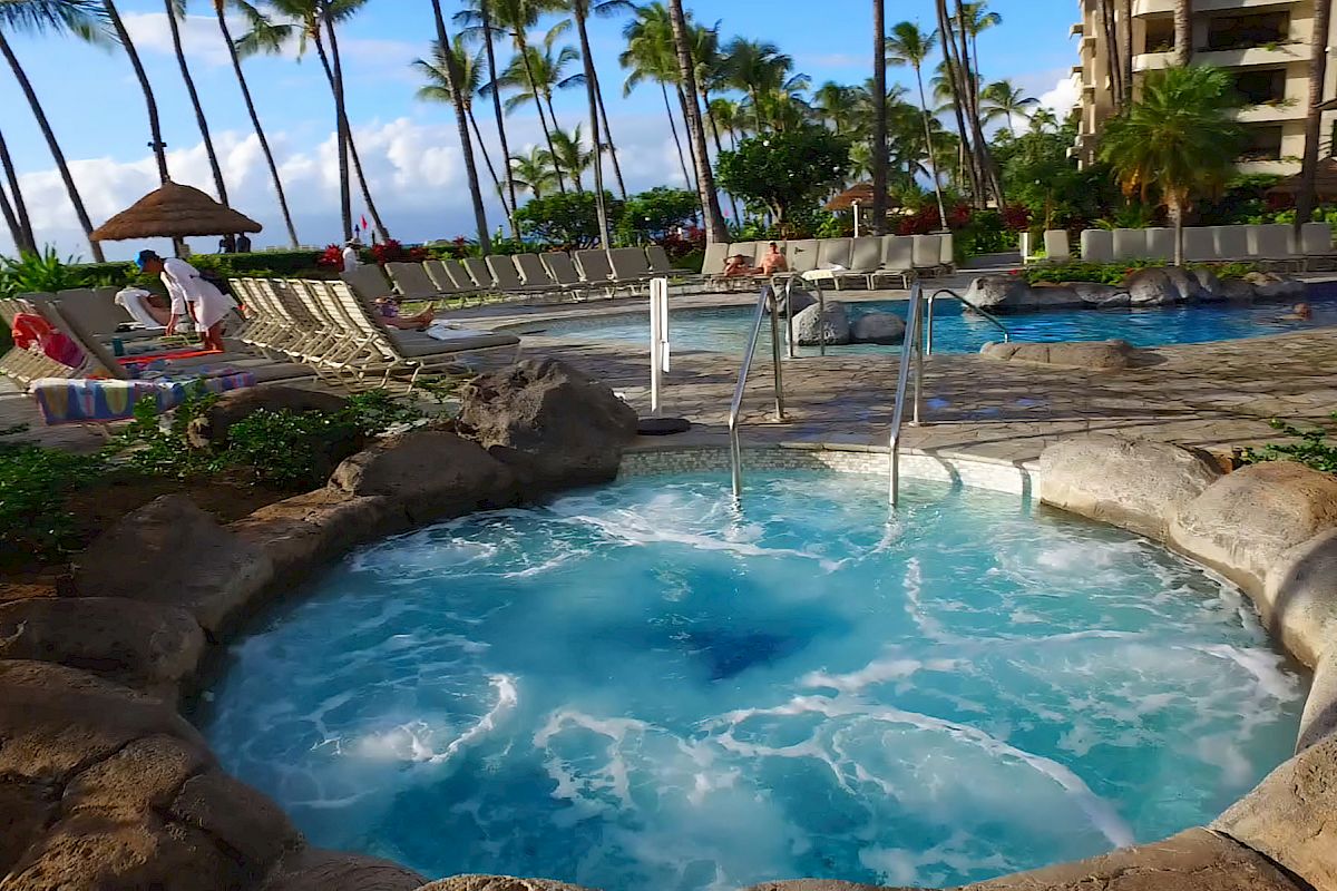 An outdoor pool area with a hot tub, sun loungers, palm trees, and people relaxing, with buildings and clear blue skies in the background.
