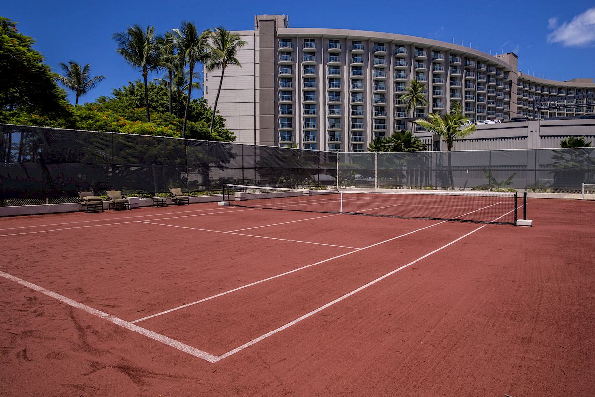 The image shows an outdoor tennis court with a clay surface, surrounded by a fence, with a tall, curved building and palm trees in the background.