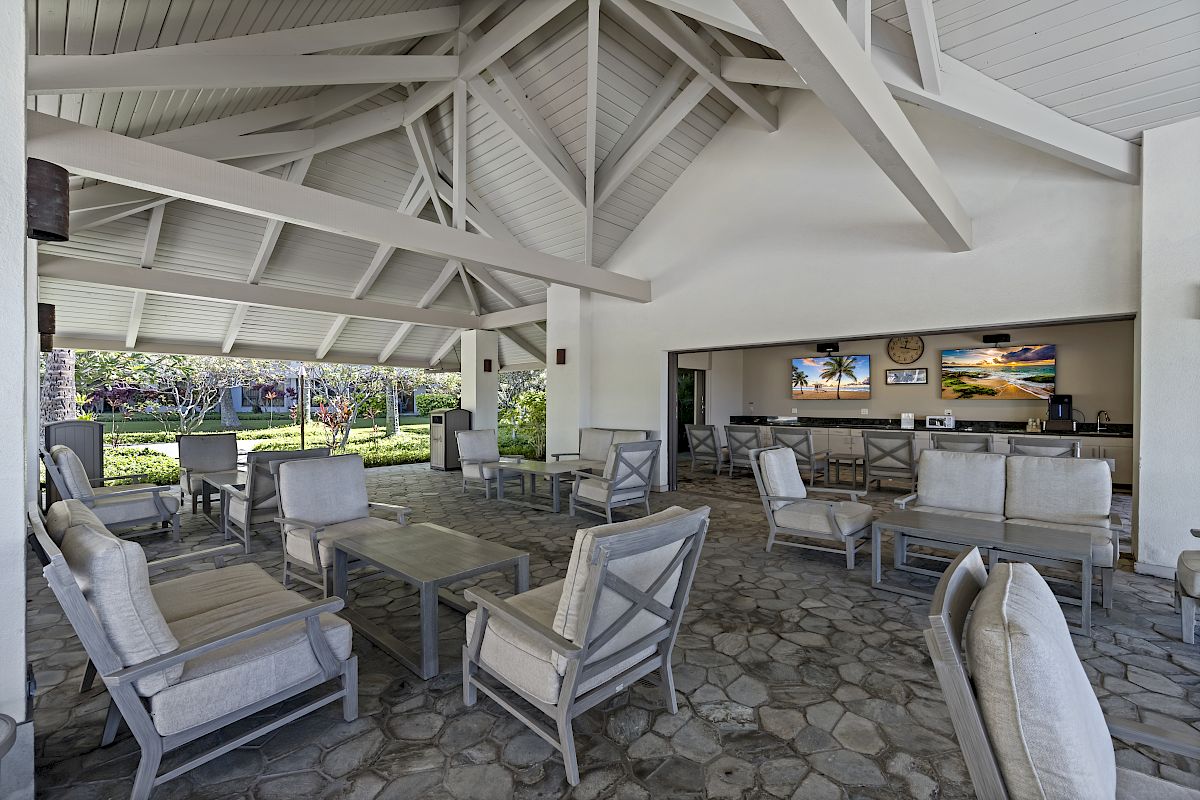 An open-air lounge area with cushioned seating, a bar counter, stone flooring, and a high beamed ceiling, overlooking a lush green outdoor space.