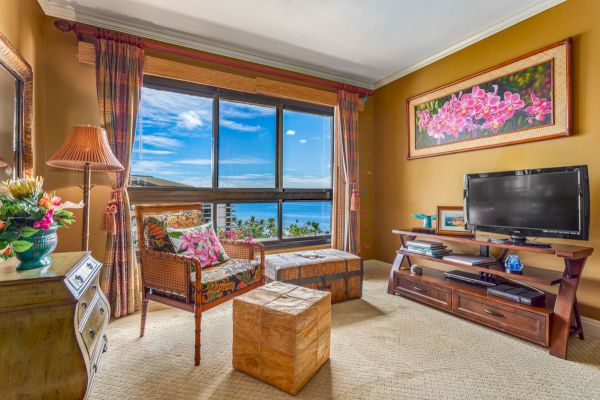 A cozy living room with a TV, wicker chair, wooden furniture, and large window with a spectacular coastal view; decorated with flowers and art.