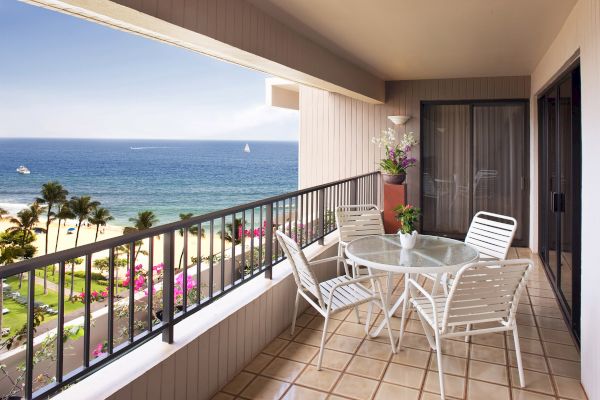 A balcony with a table and four chairs overlooks a scenic beach and ocean view, complete with palm trees and beachfront greenery.