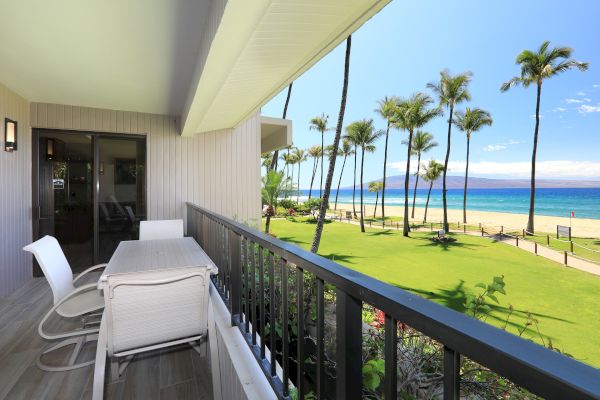 A balcony with white furniture overlooks a lush green lawn, tall palm trees, and a beautiful beach with clear blue water and a sunny sky.