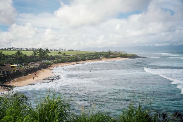 A beachscape with waves crashing on the shore, a few houses, greenery, and hills in the background under a partly cloudy sky ending the sentence.