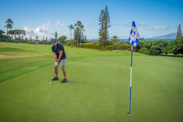 A person is putting on a golf course with a blue and white checkered flag in the foreground, surrounded by greenery and palm trees.