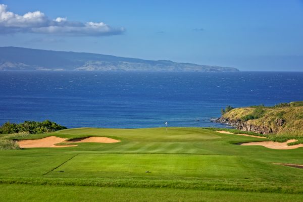 A scenic golf course by the ocean, with clear blue skies and distant mountains visible across the water, featuring lush green fairways and sand bunkers.