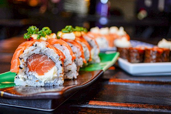 The image shows a plate of sushi rolls topped with sliced salmon and green onions, placed on a dark serving platter.