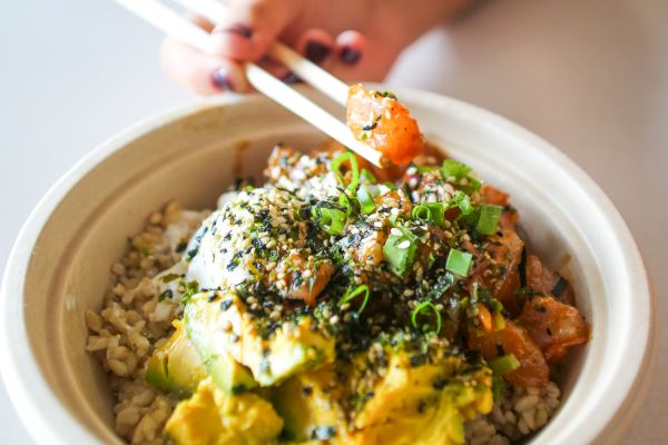 A hand holding chopsticks is reaching for a poke bowl, which includes rice, avocado, salmon, and toppings like sesame seeds and seaweed.