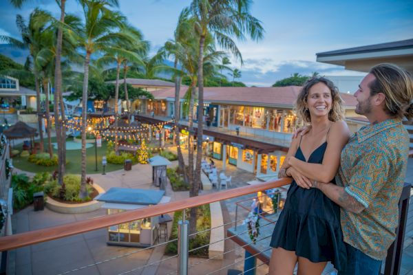 A couple smiles while overlooking a lively shopping center with palm trees and shops lit for the evening.