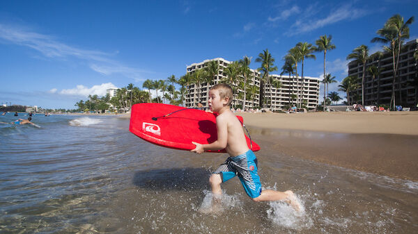 A child holding a red bodyboard runs through shallow water on a sunny beach with buildings and palm trees in the background.