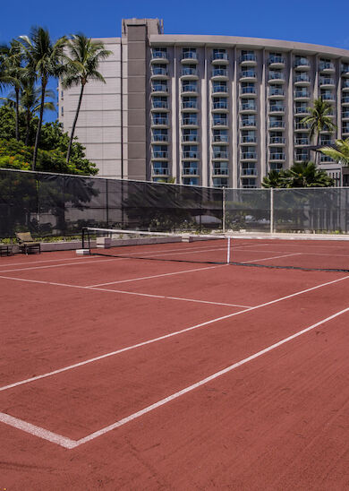 The image shows a red clay tennis court with a net and a high-rise building in the background, surrounded by tropical trees and clear blue skies.