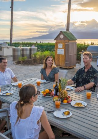 A group of six people is enjoying a meal outdoors at a beachside location with a scenic sunset in the background, while a chef cooks on a grill.