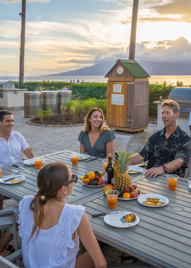 A group of six people is enjoying a meal outdoors at a beachside location with a scenic sunset in the background, while a chef cooks on a grill.