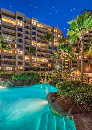 The image shows a nighttime view of a resort with a large illuminated swimming pool, surrounded by palm trees, while high-rise buildings stand in the background.