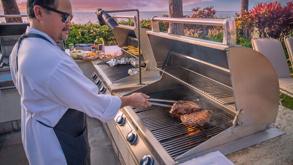 A person wearing glasses and an apron is grilling steaks on an outdoor barbecue with scenic surroundings at sunset, holding tongs for cooking.