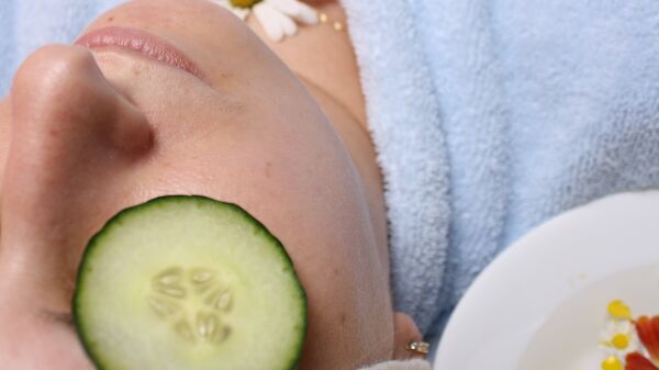 A person is relaxing with a cucumber slice on their eye, wearing a blue towel, and a flower on their chest. A plate of fruits is nearby.