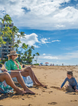 A family relaxes on a sunny beach with a child playing in the sand, while a building and palm trees are visible in the background.