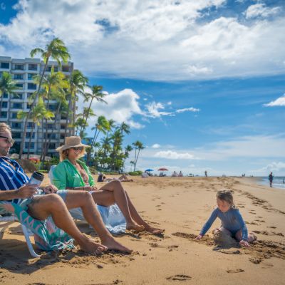 A family relaxes on a sunny beach with a child playing in the sand, while a building and palm trees are visible in the background.