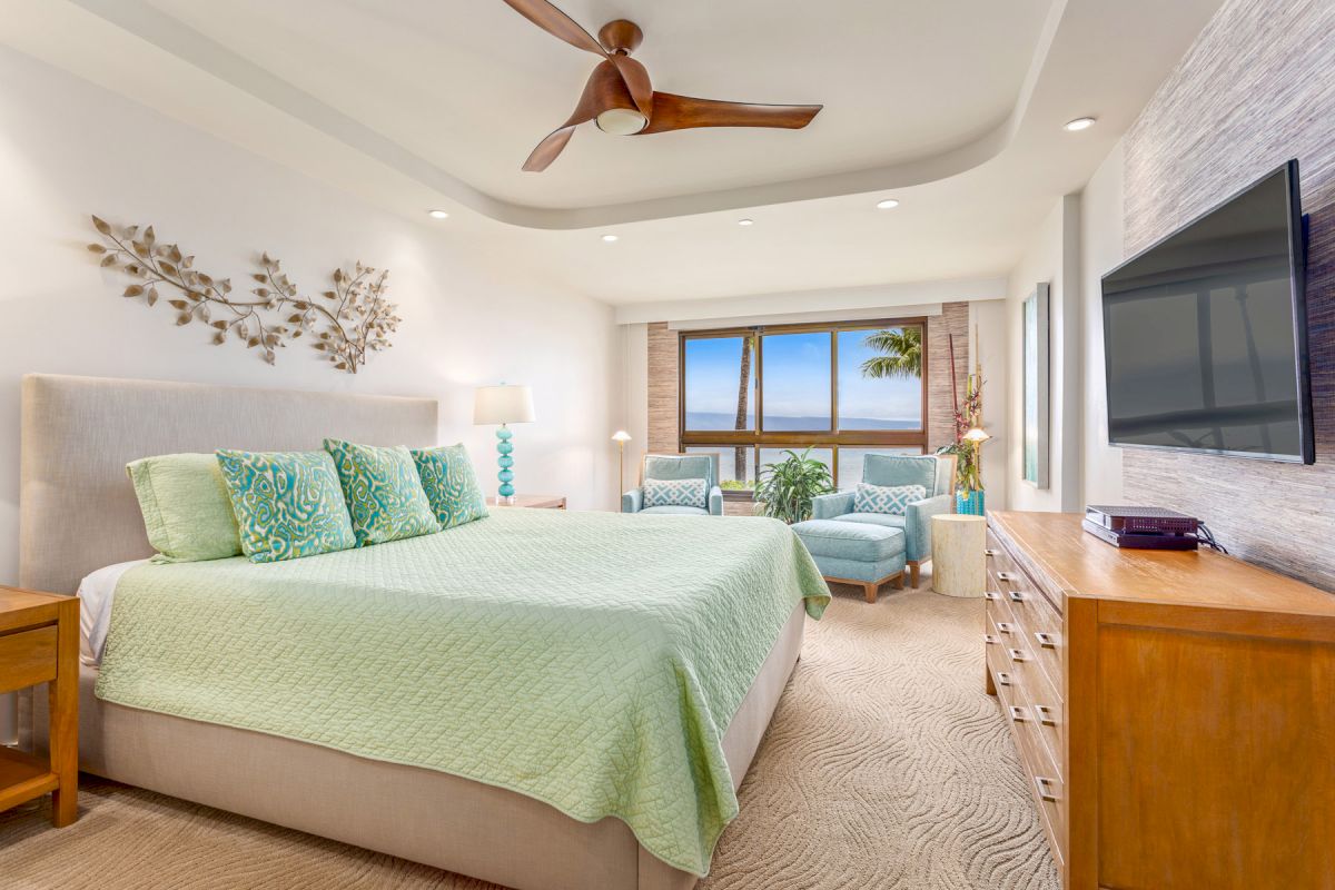 A modern bedroom with a large bed, green bedding, and matching pillows, featuring a TV, wooden furniture, and a seating area near the window.