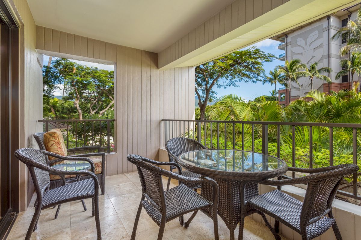 The image shows a balcony with a round glass table and four chairs. It's overlooking a lush tropical garden with palm trees and other greenery.