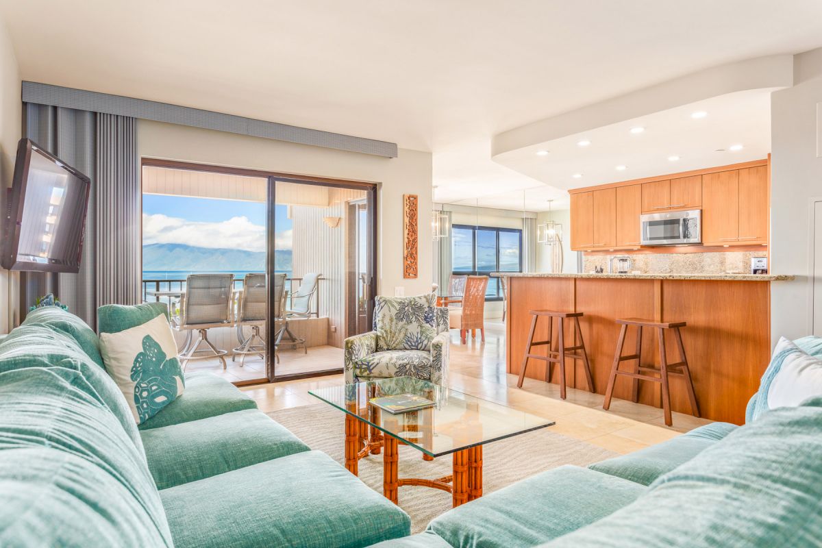 A modern living room with teal furniture, a glass coffee table, a mounted TV, and a kitchen area with wooden cabinets, overlooking a scenic ocean view.