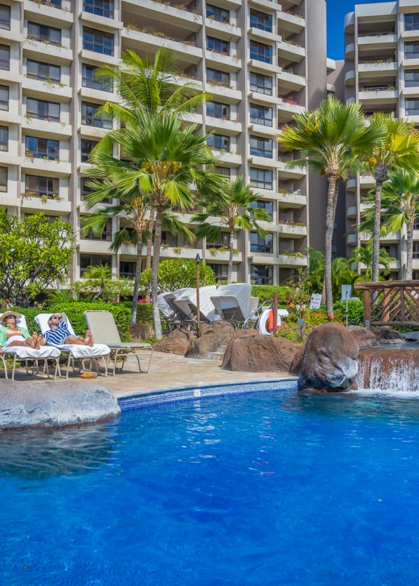 A serene pool area with lounge chairs, people relaxing, palm trees, and a multi-story building in the background.