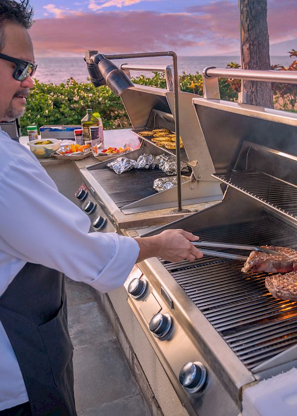 A person wearing sunglasses and an apron is grilling meat on a barbecue near a scenic outdoor setting with a sunset in the background.