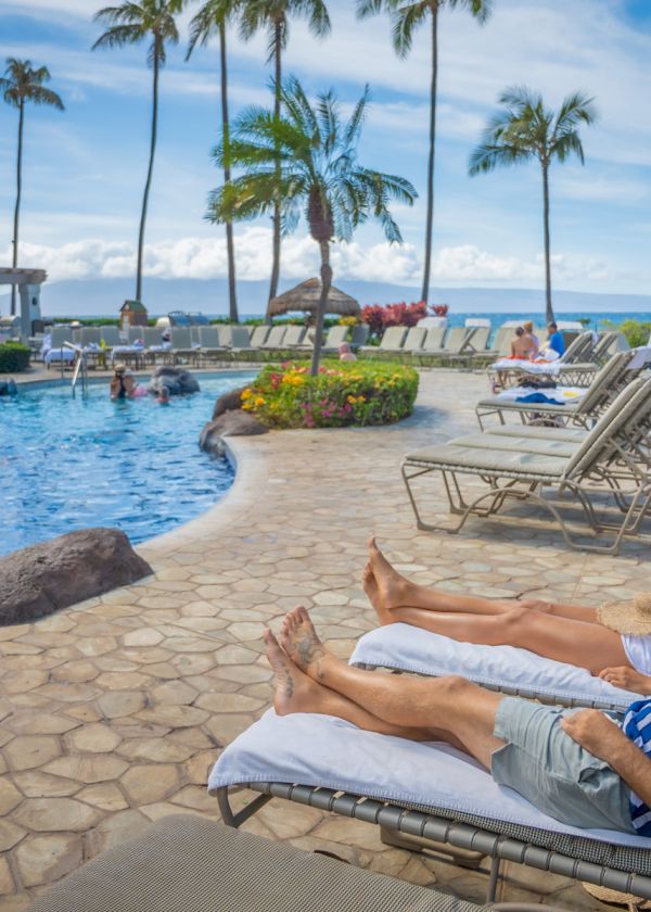 People are lounging by a pool at a tropical resort with palm trees and blue skies in the background, enjoying a relaxing day.