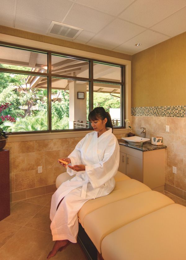 A person in a white robe is sitting in a spa room, comfortably reading something, surrounded by a tranquil, well-lit environment with a massage table.