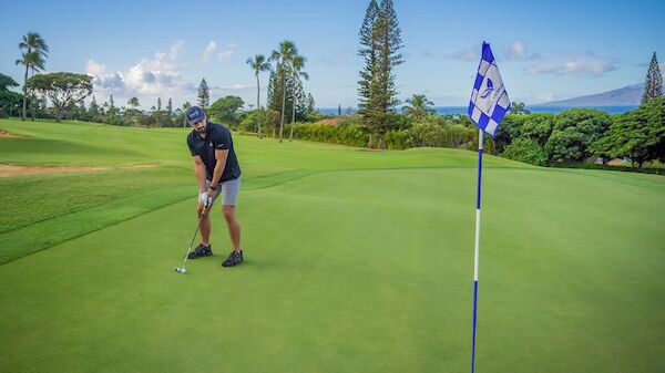 A person is putting on a green golf course with a flag marked "18," surrounded by trees, and a scenic background of a mountain and blue sky.