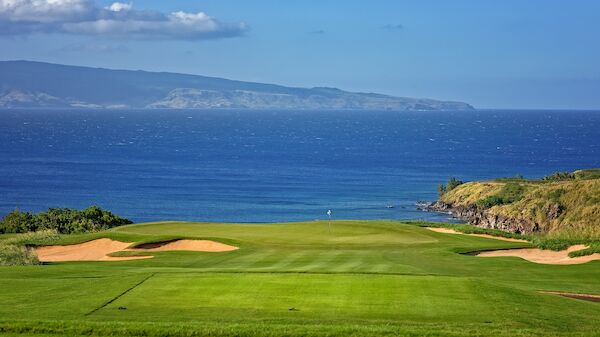 A green golf course with sand bunkers, overlooking a blue ocean, and an island in the background, under a clear sky with scattered clouds.