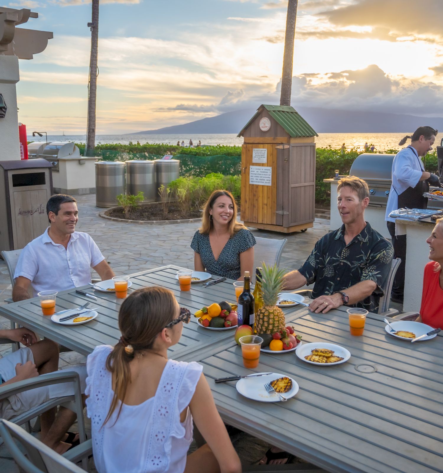 A group of six people are sitting at an outdoor table enjoying a meal with drinks, fruit, and sunset views while someone cooks at a grill in the background.