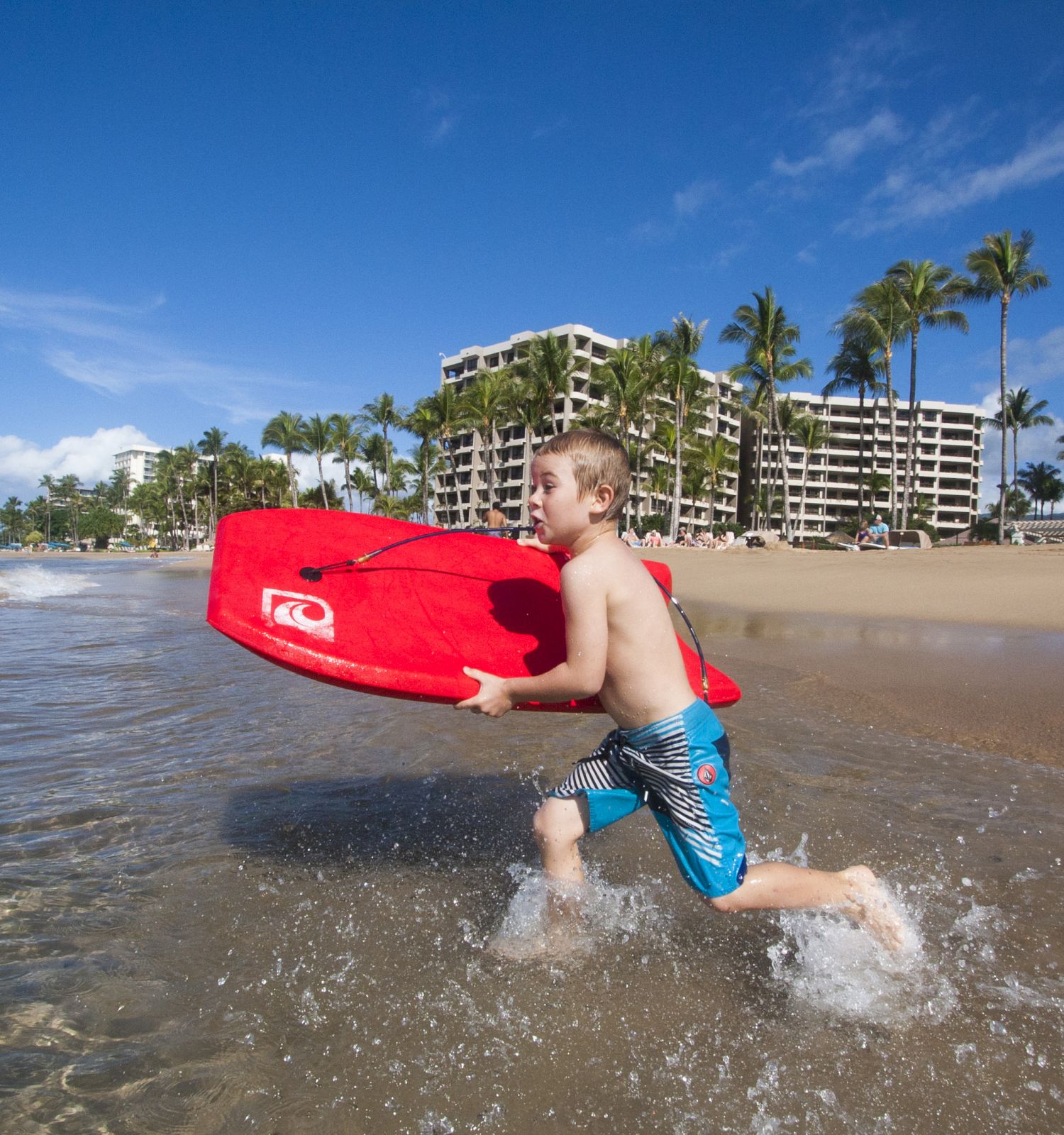 A child holding a red boogie board is running along the beach with the ocean in the background, near a line of palm trees and beachfront buildings.