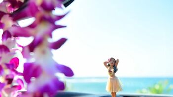 A hula dancer figurine on a car dashboard with an ocean view in the background, and blurred purple flowers in the foreground.