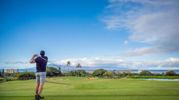 A man is playing golf on a lush green golf course under a bright blue sky with scattered clouds and a scenic background.