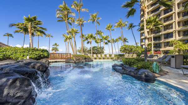 An inviting tropical pool area features palm trees, lounge chairs, a waterfall, and a bridge against a backdrop of a multi-story building and blue sky.